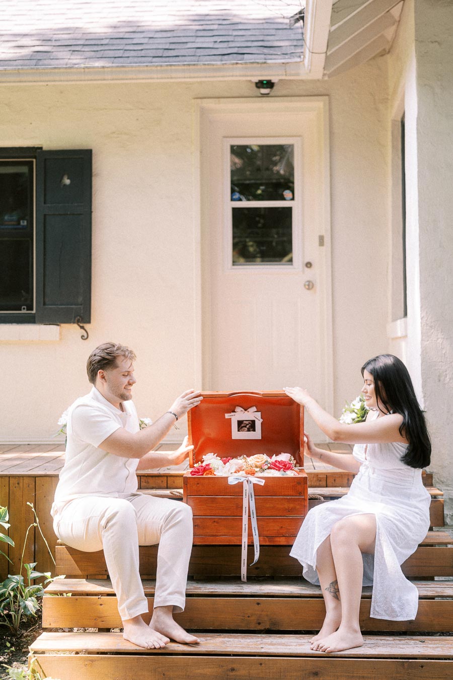 Couple in white outfits opening a wooden box on porch steps.