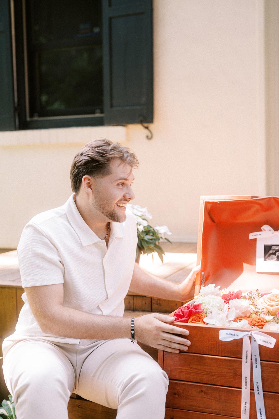 Man in white sitting beside an open wooden box filled with flowers.