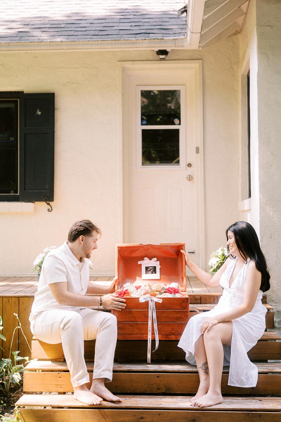 Couple in white outfits sitting on porch steps with an open memory box.
