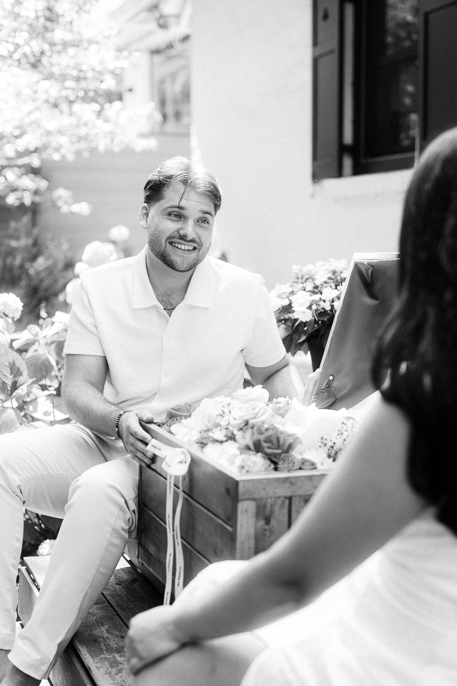 Black and white engagement photo of a smiling man seated across from his partner outdoors.