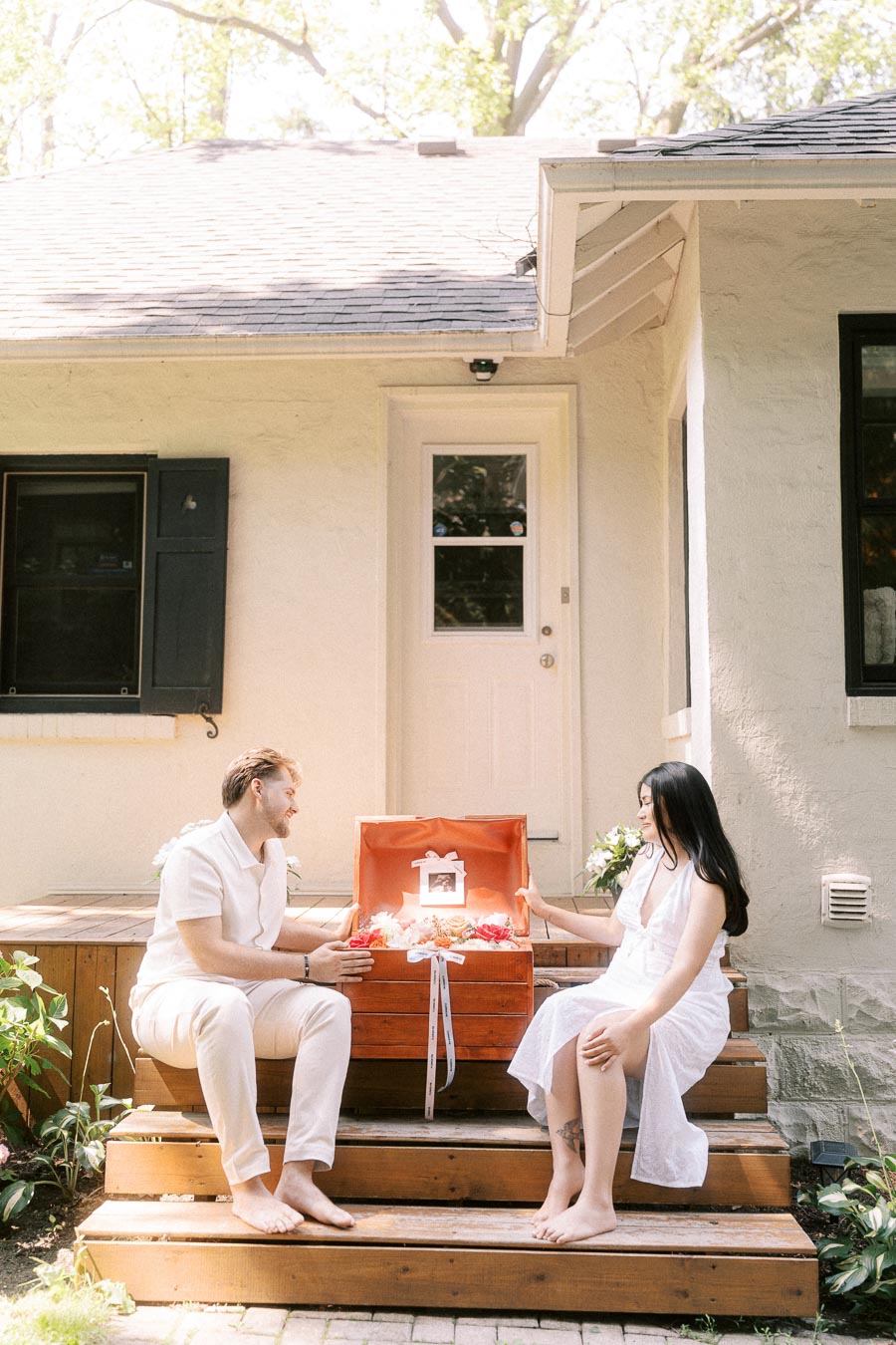 Engaged couple in white outfits sitting on porch steps with an open gift box.