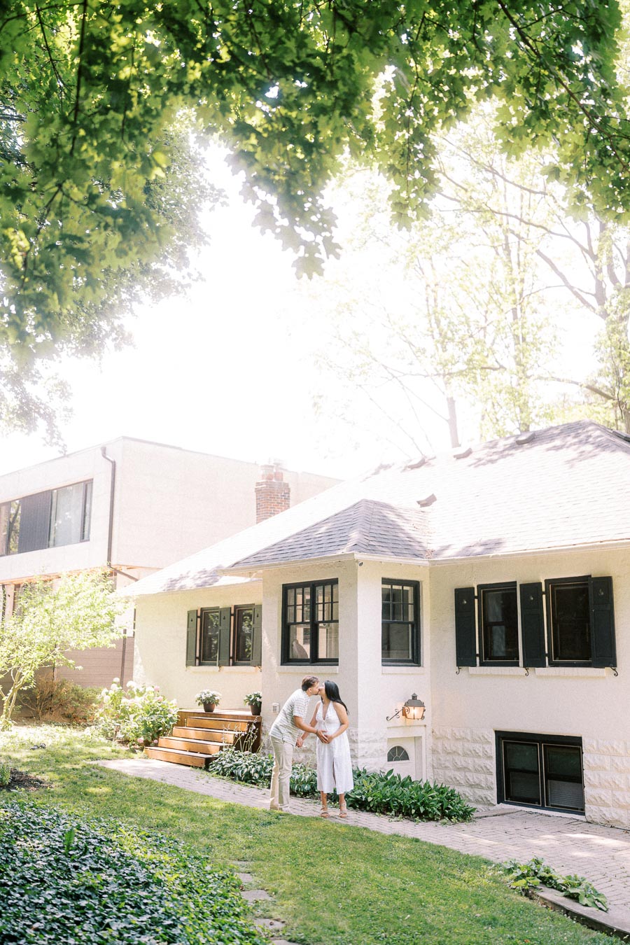 Engaged couple kissing outside a white house surrounded by leafy greenery.