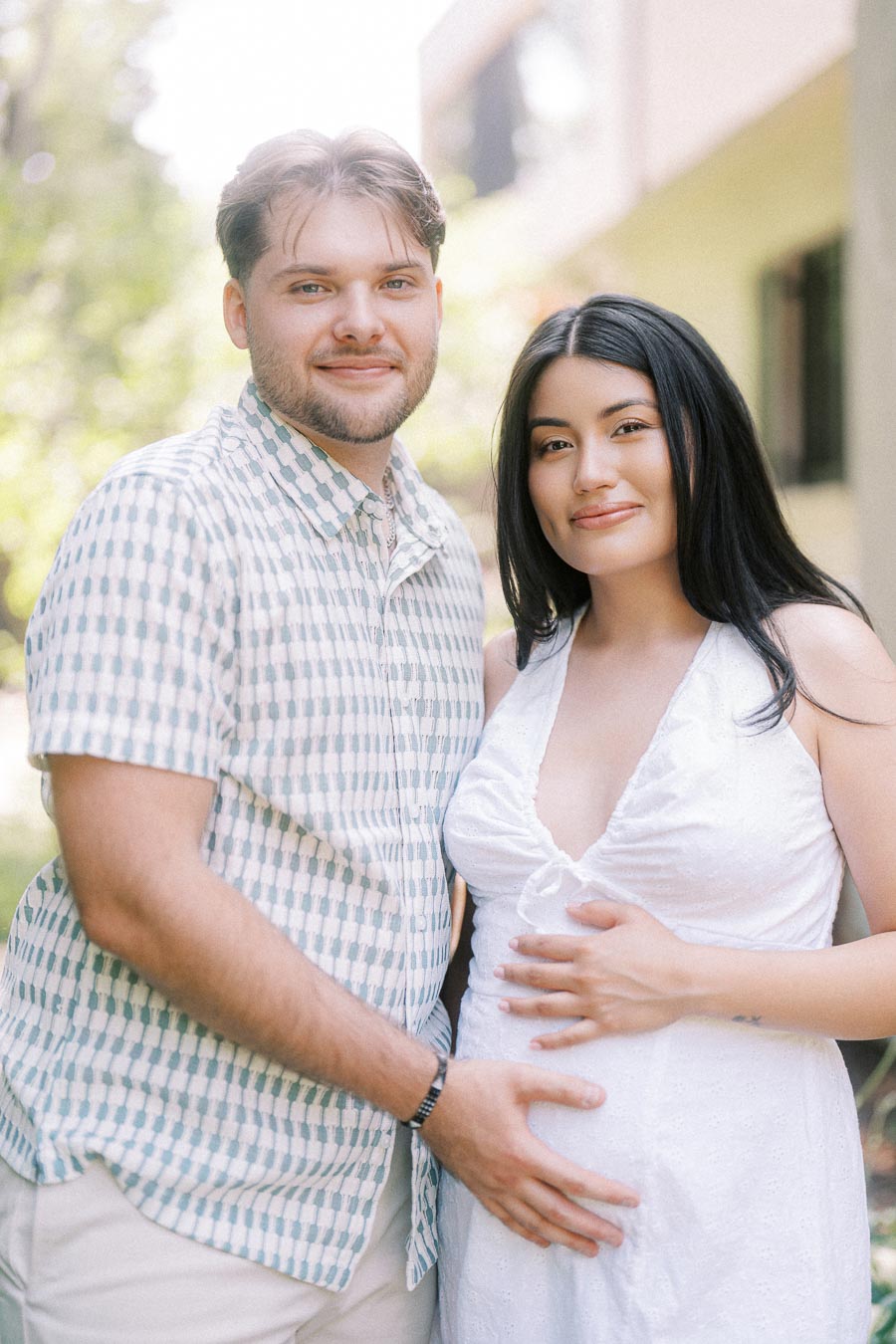 Couple posing outdoors, both hands resting on the woman’s pregnant belly.