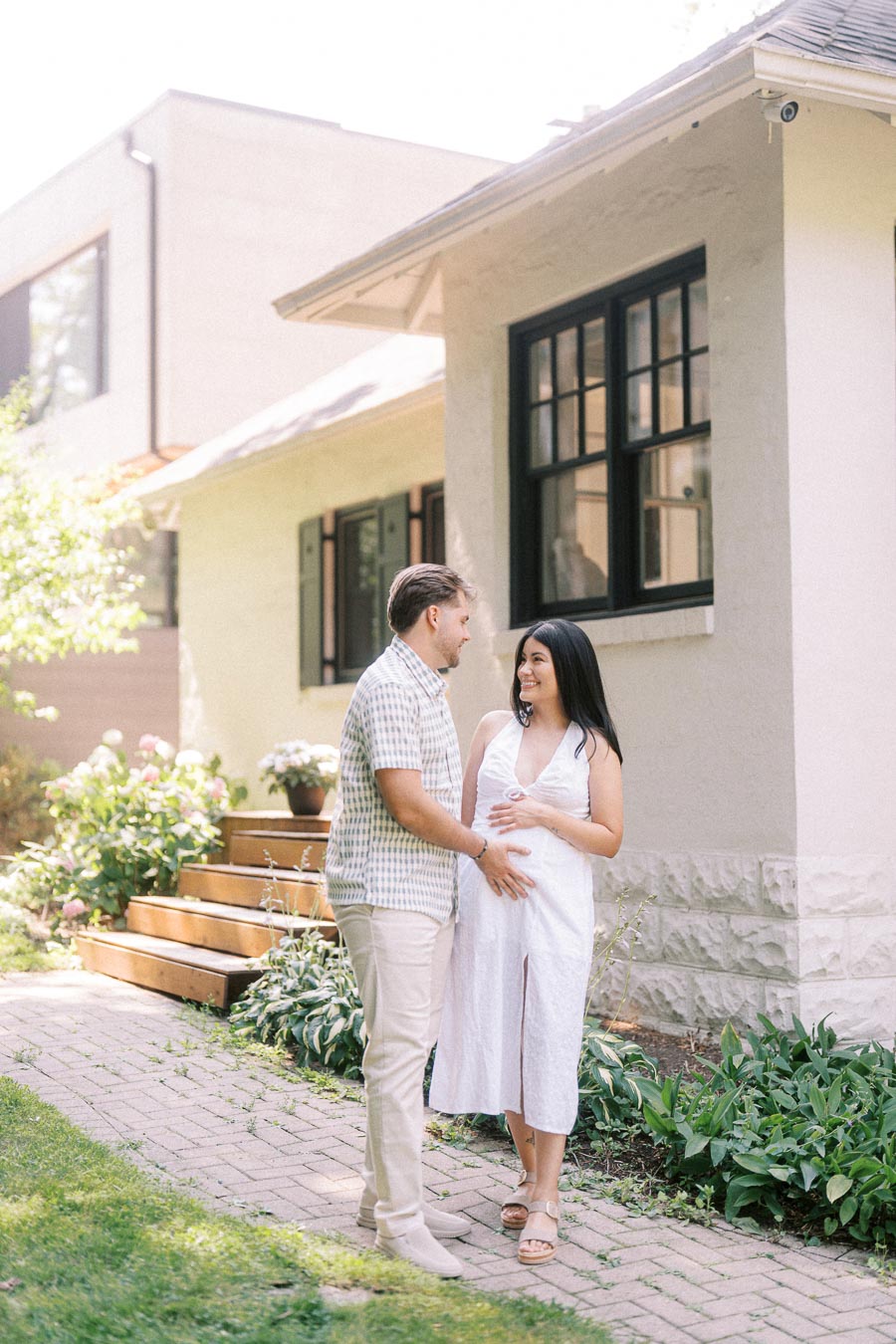 Expectant couple smiling together beside a garden walkway and house.