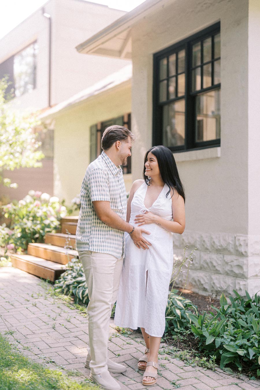 Couple smiling together outside a house, with hands resting on her pregnant belly.
