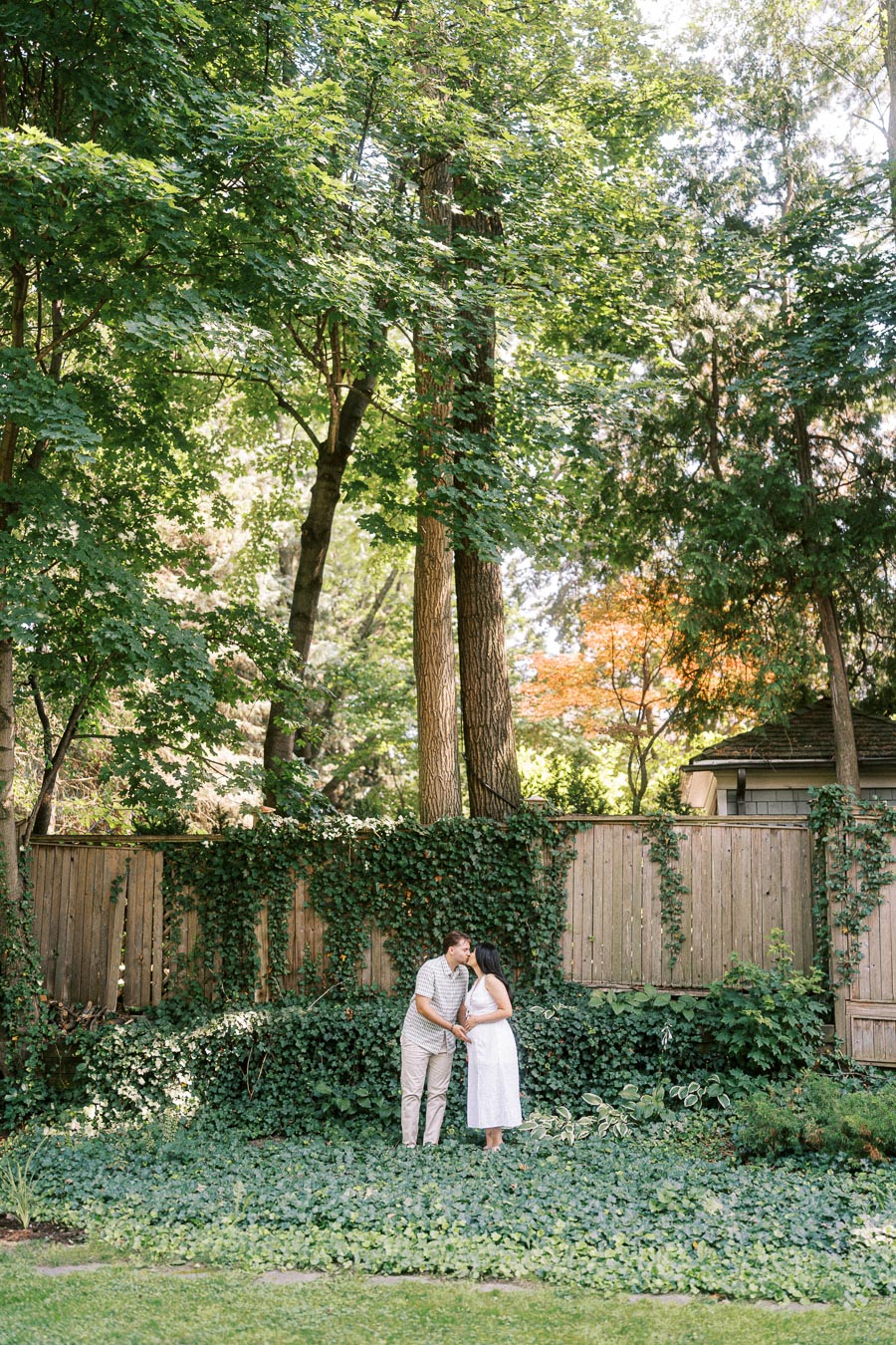 Engaged couple holding hands in a leafy garden beneath tall trees.