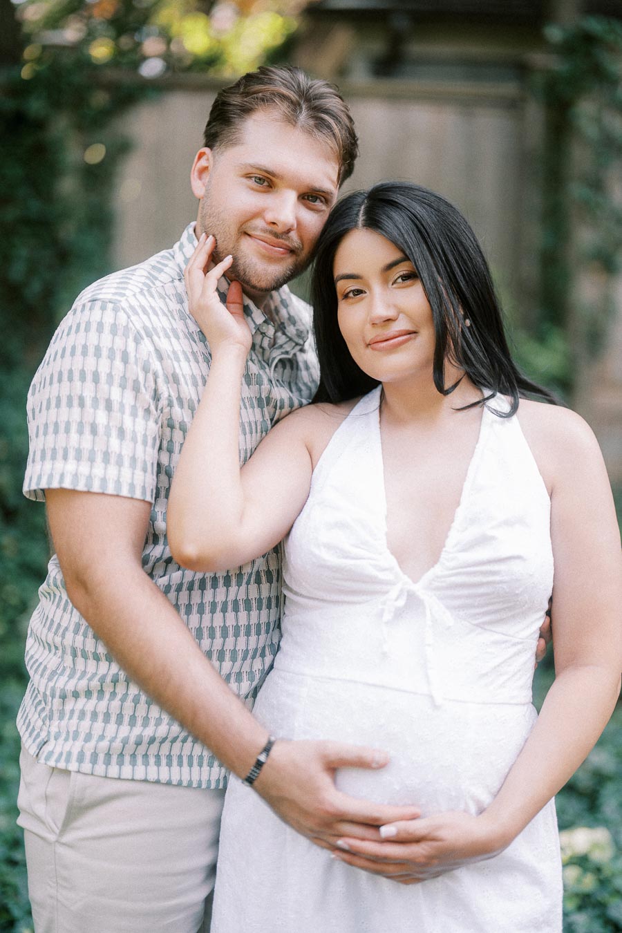 Expectant couple posing closely in a leafy garden setting during an engagement photo.
