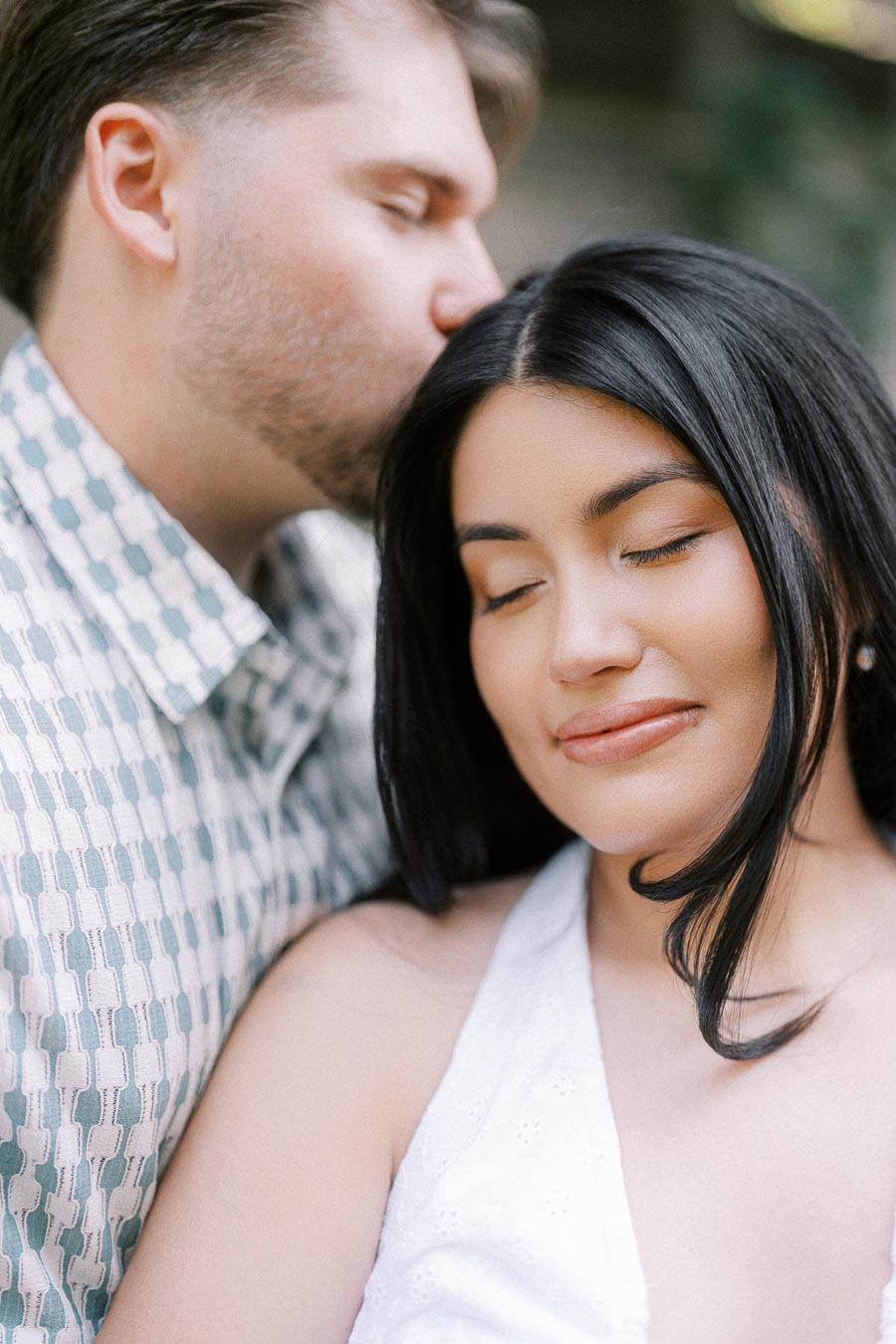 Close-up engagement portrait of a man kissing his partner’s forehead outdoors.