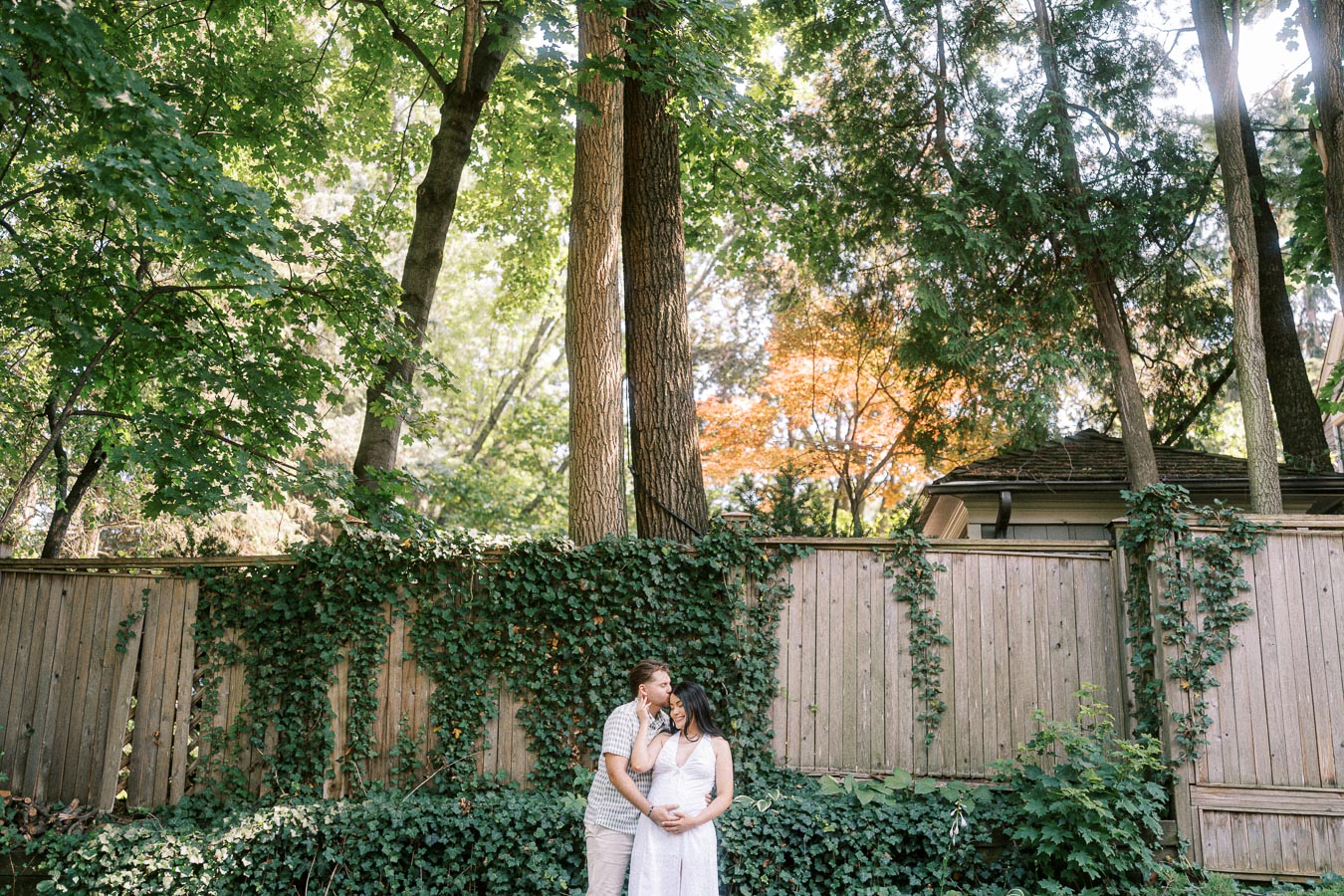 Couple embracing beside an ivy-covered fence in a leafy Toronto setting.
