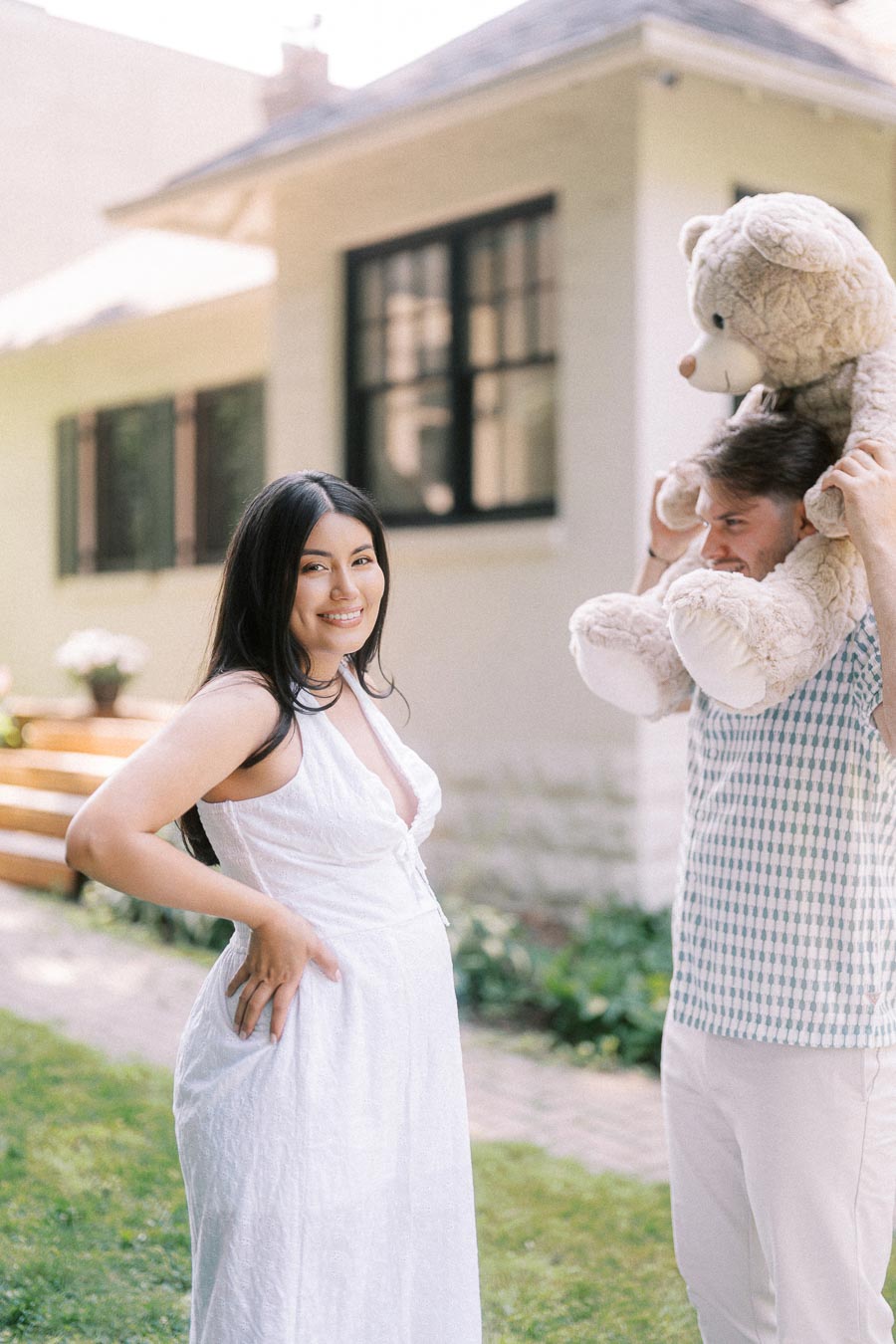 Expectant couple outside a house, with a man holding a large teddy bear.