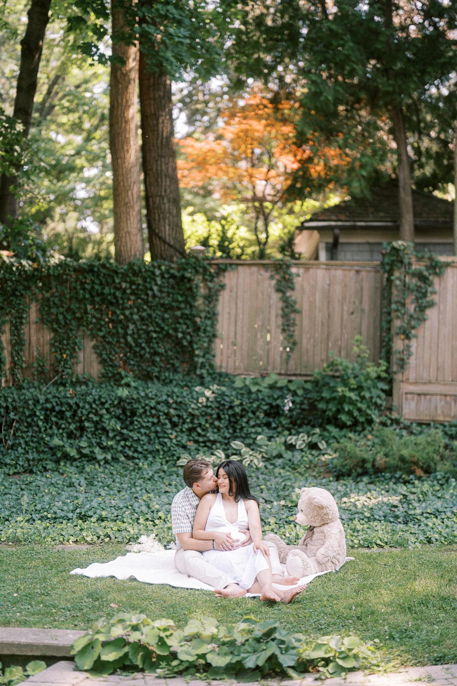 Expectant couple sitting on a blanket in a leafy Toronto garden with a teddy bear.