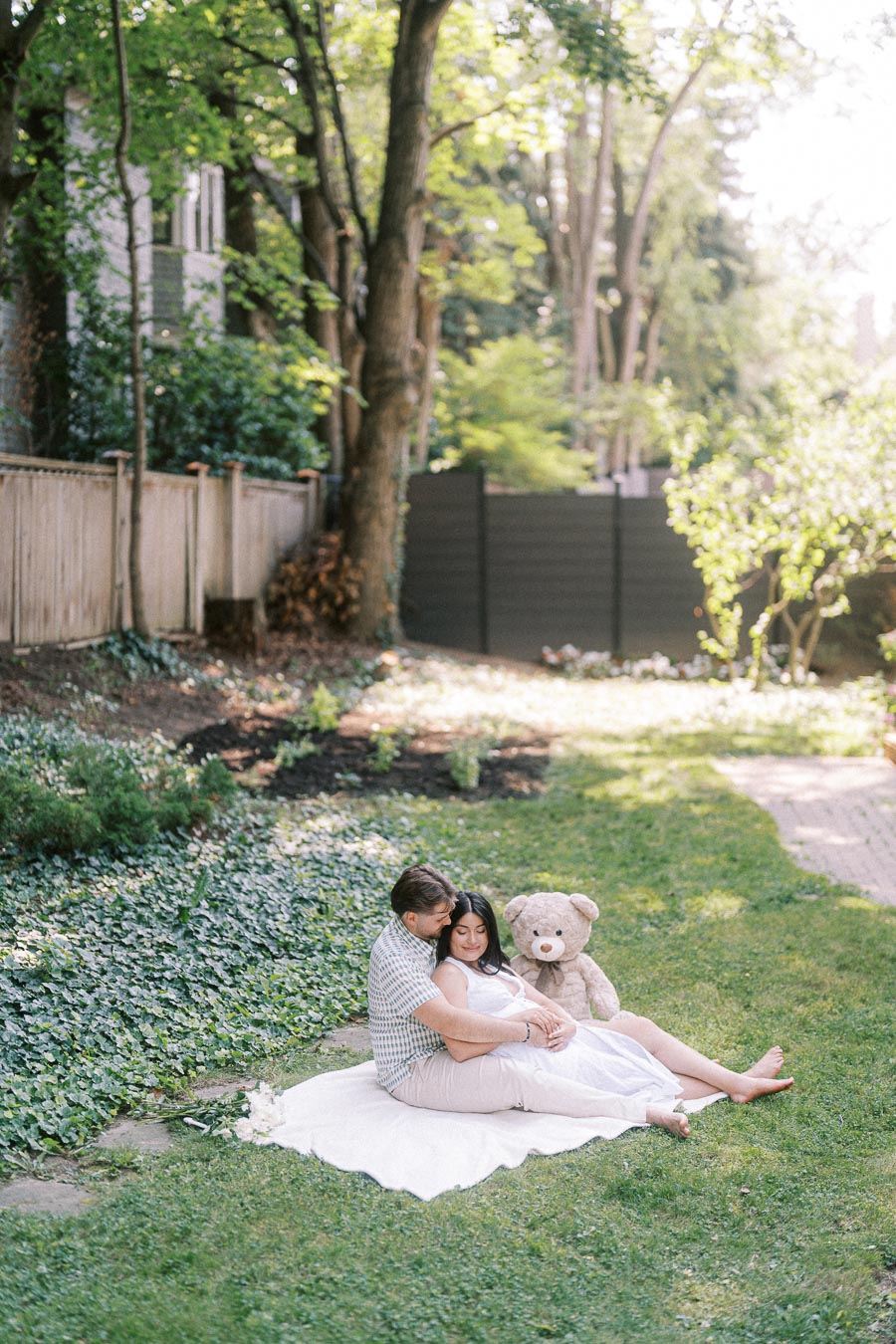 Expectant couple sitting on a blanket in a garden with a teddy bear.