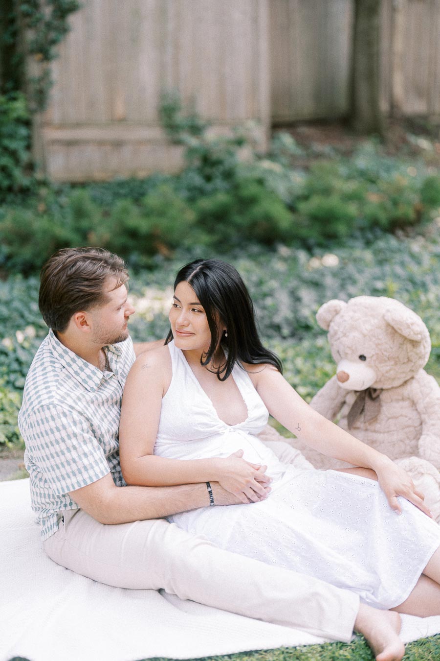 Expecting couple sitting on a blanket in a garden with a teddy bear nearby.