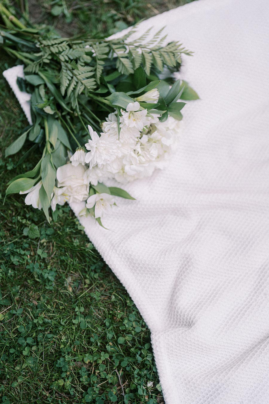 White bouquet with greenery resting on a textured blanket in the grass.