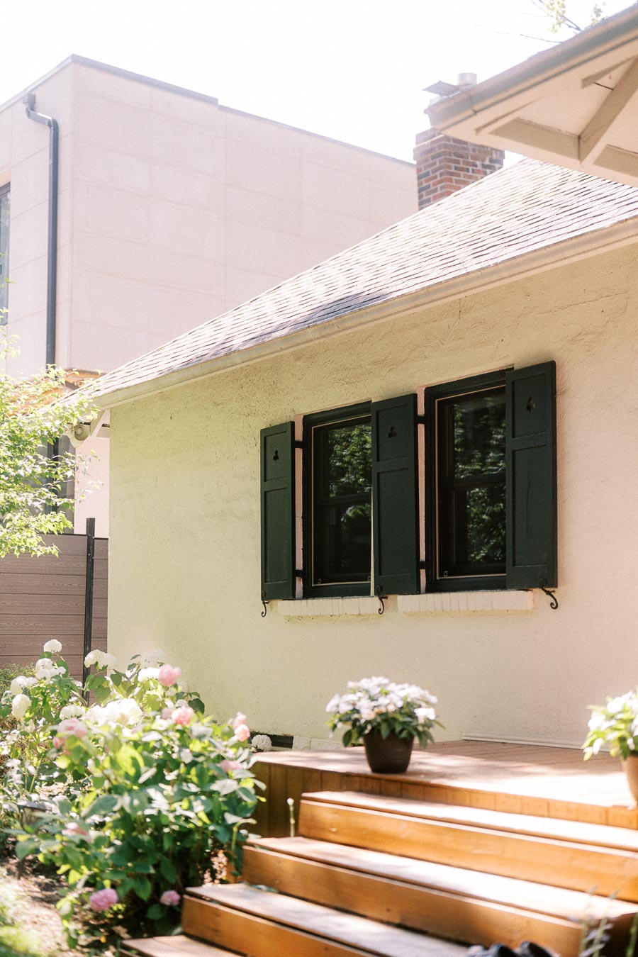 Sunlit house exterior with green shutters, wooden steps, and garden flowers.