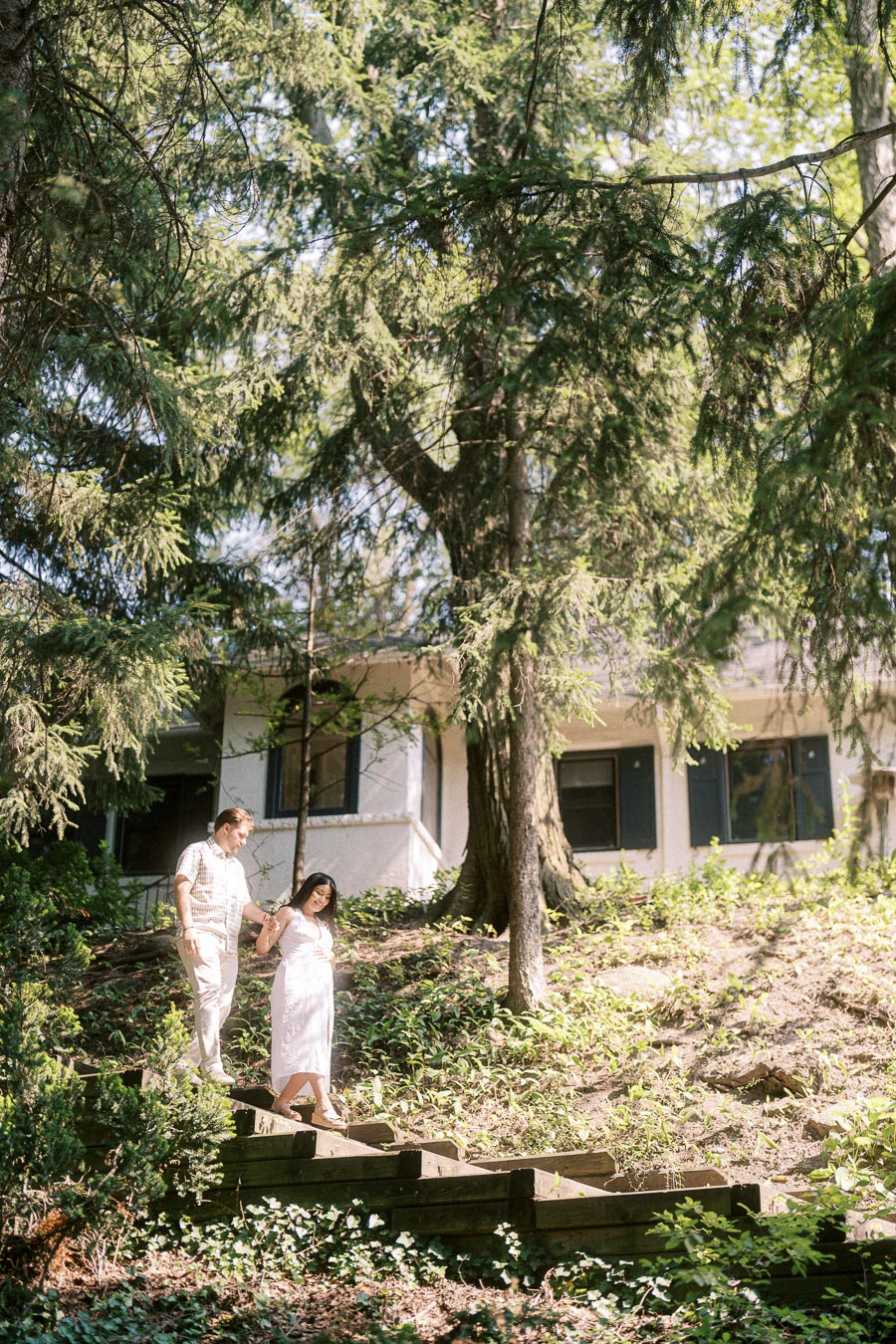 Engaged couple holding hands on wooden steps beneath tall evergreen trees.