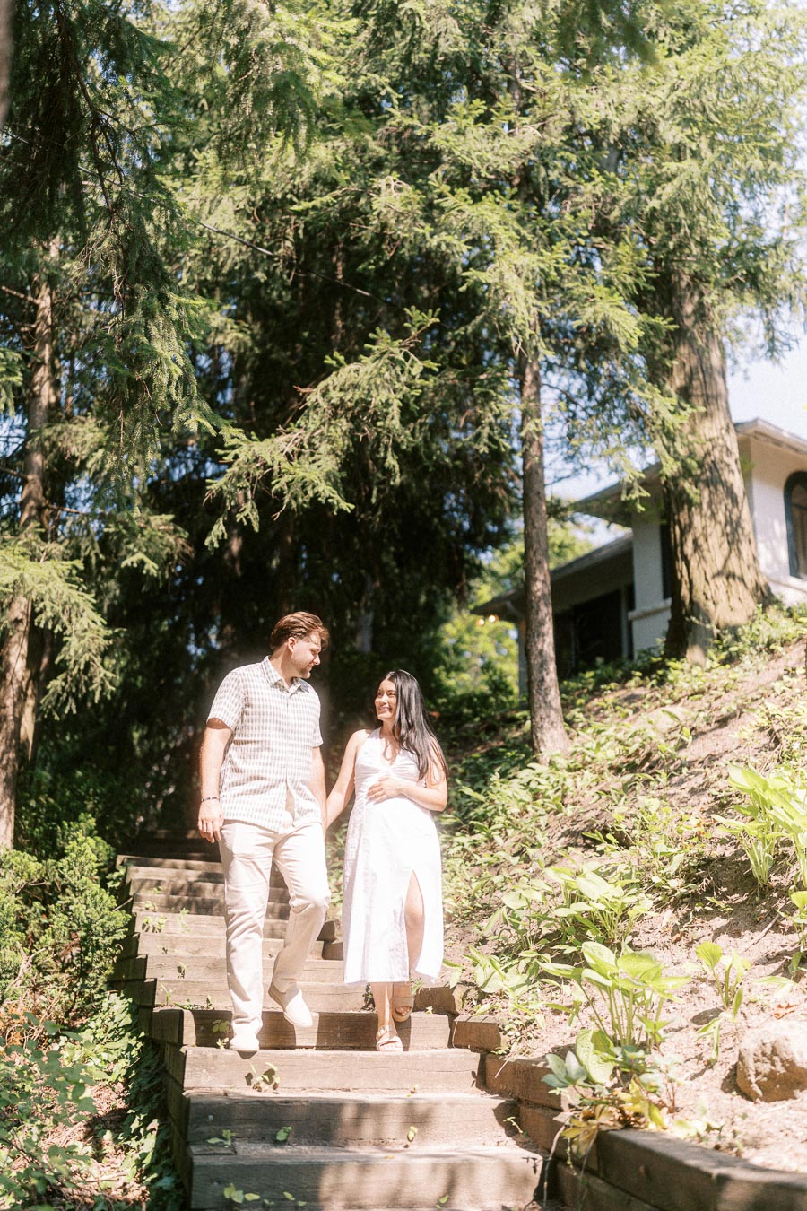 Engaged couple walking down outdoor wooden steps beneath tall evergreen trees.