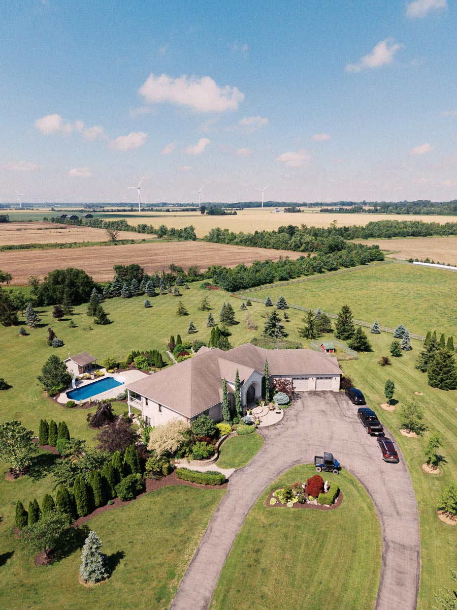 Aerial view of a countryside home with gardens, pool, driveway, and surrounding fields.