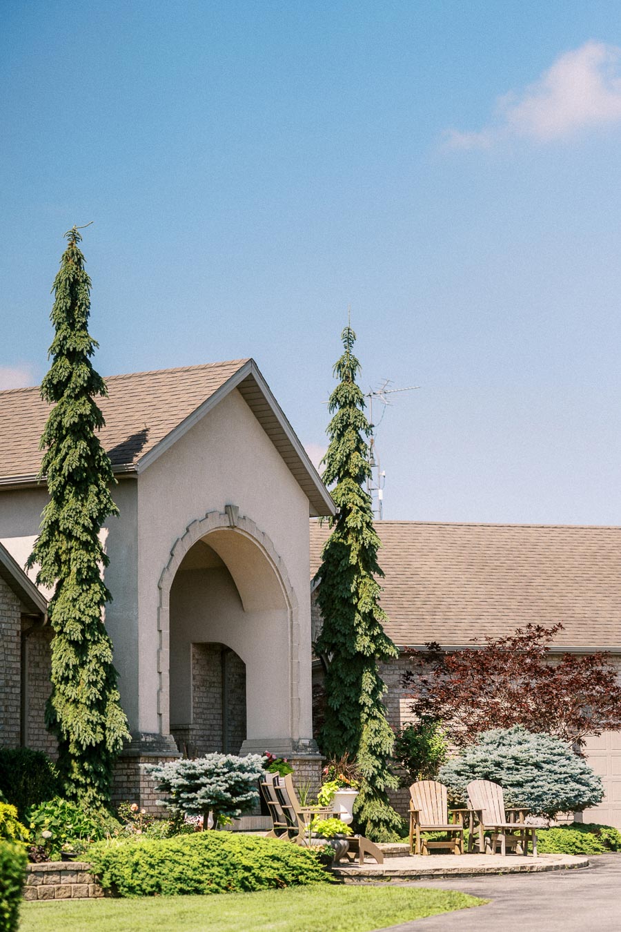 Landscaped home entrance with tall evergreens, arched doorway, and wooden chairs on a patio