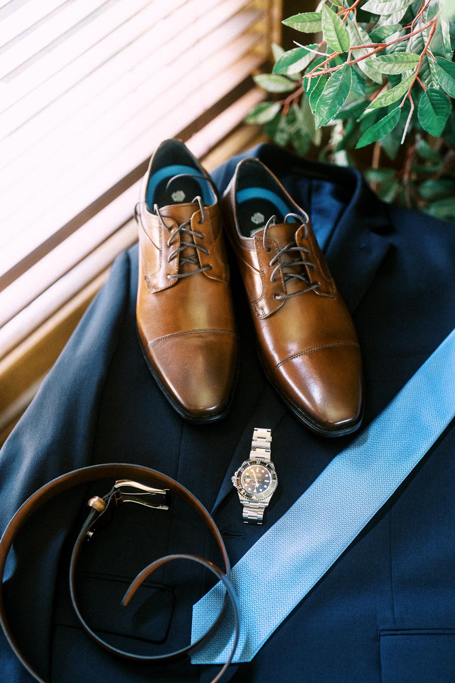 Brown dress shoes, blue tie, belt, and watch arranged on a navy suit.