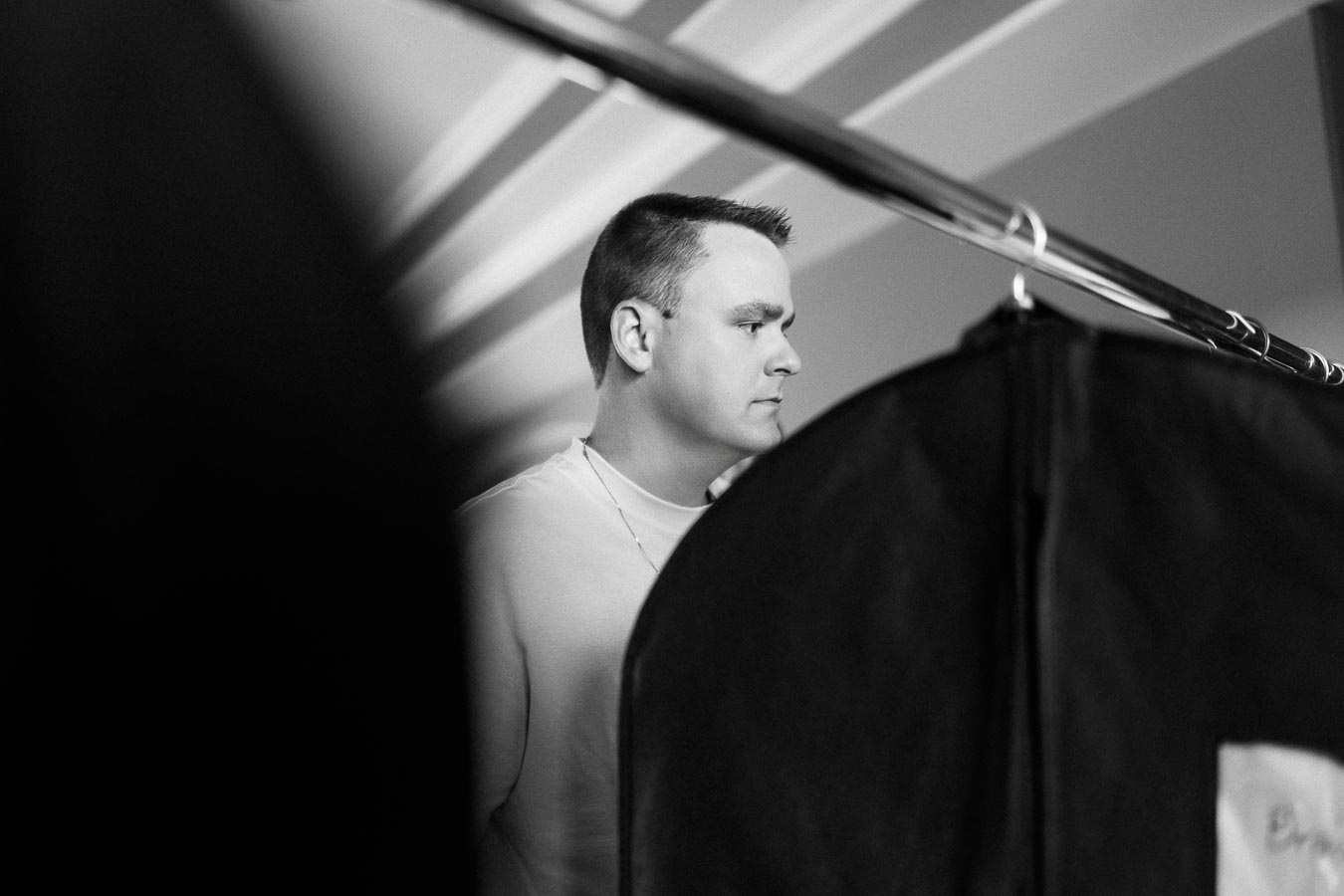Man in a white T-shirt stands behind garment bags during indoor pre-wedding preparations.