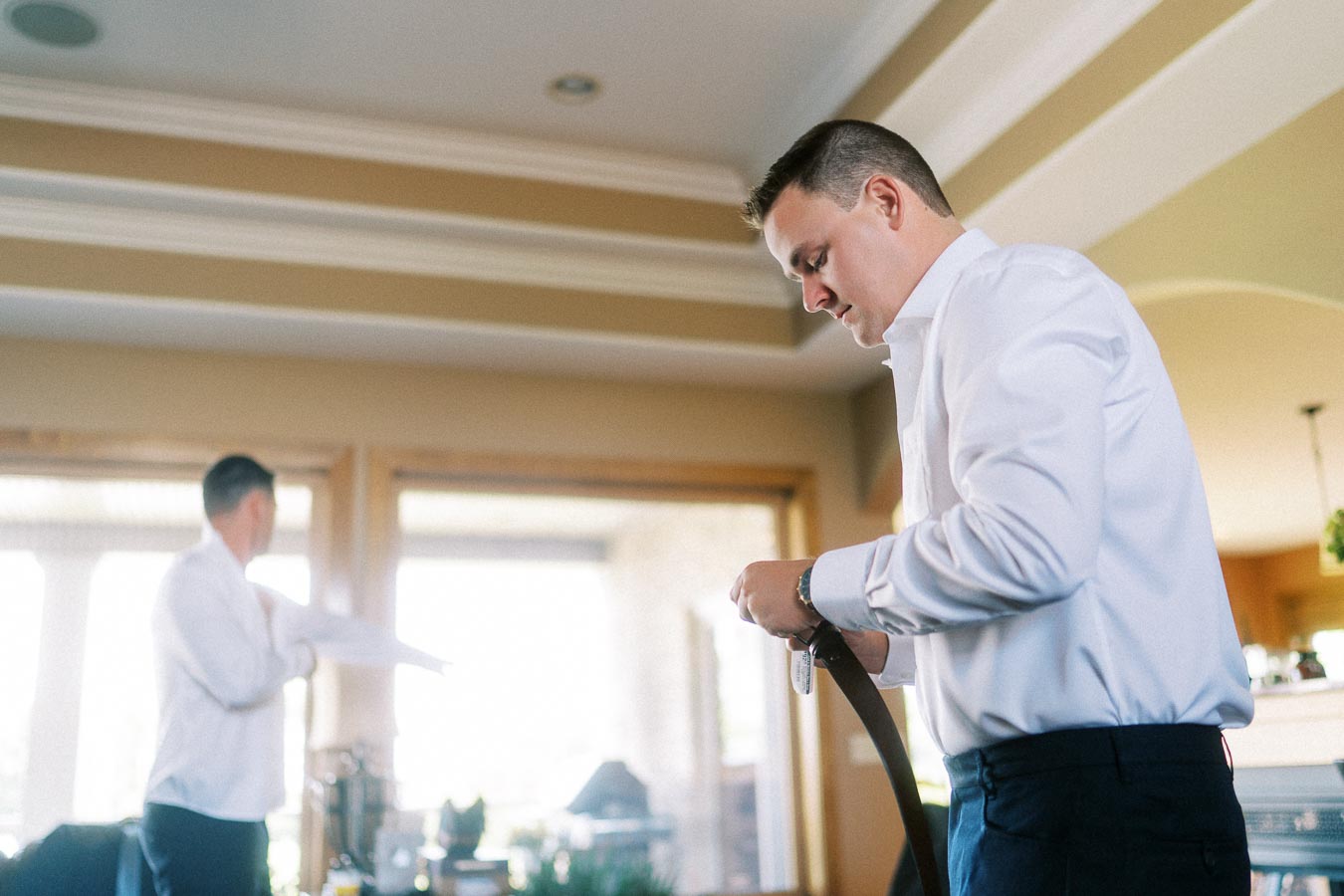 Groom in a white shirt fastening his belt at home before the wedding.