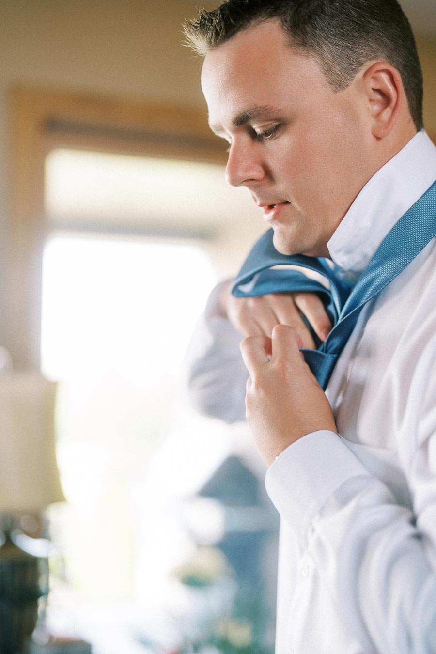 Man in a white dress shirt adjusting a blue tie indoors near soft window light.