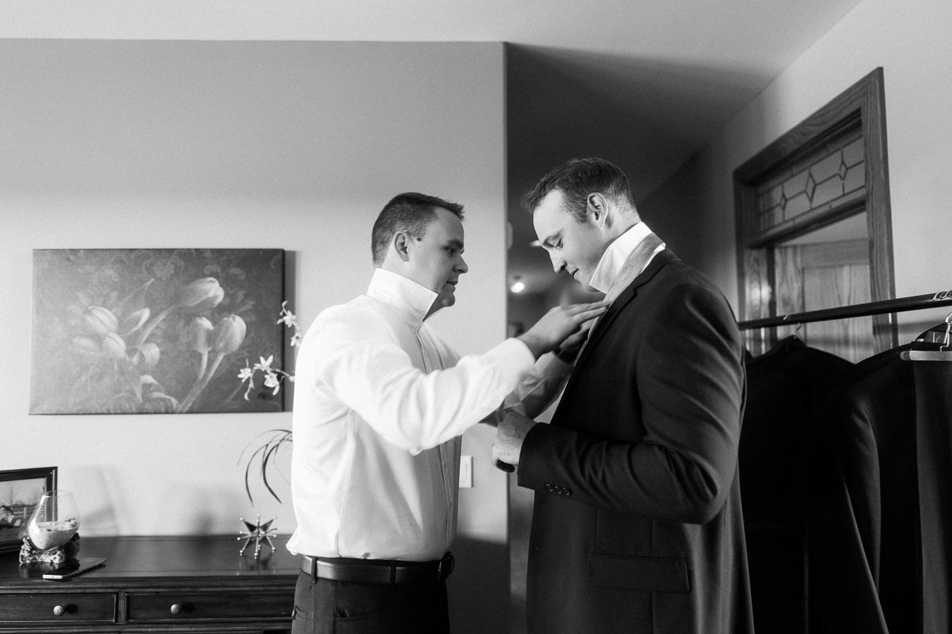 Man helps groom adjust his suit jacket indoors before the wedding.