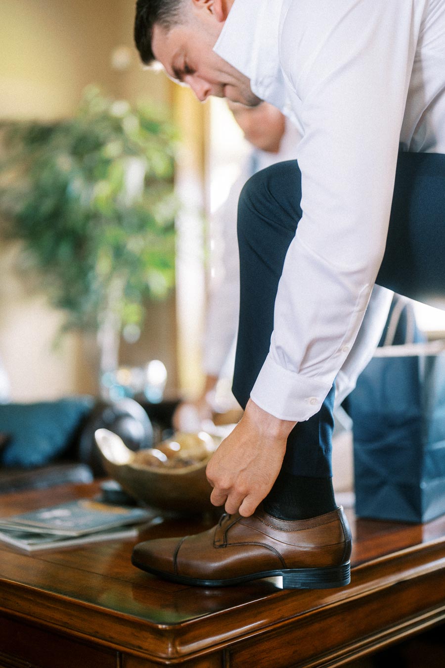 Groom in a white shirt tying brown dress shoes indoors before the wedding.