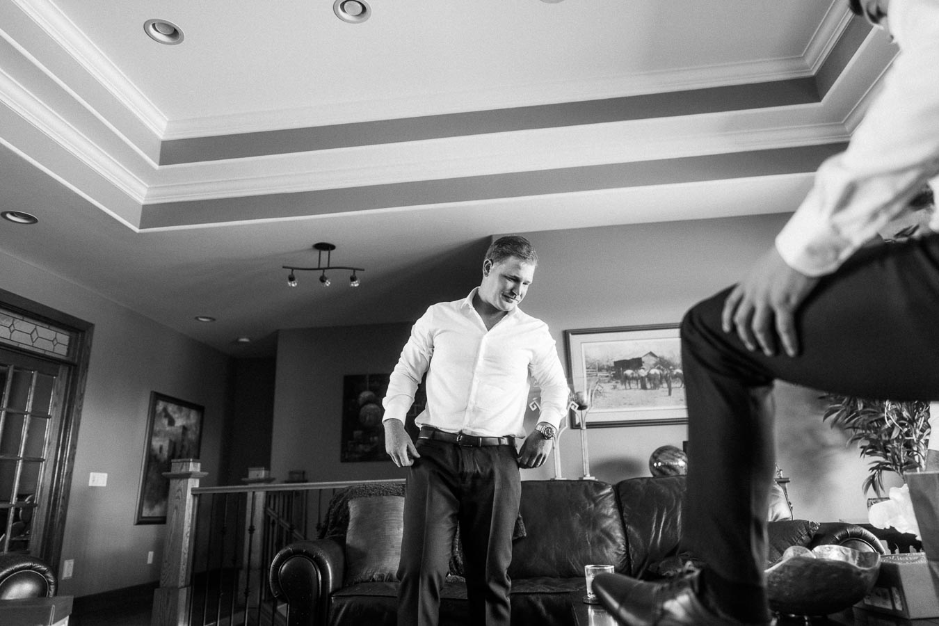 Man in a white dress shirt adjusts his trousers in a living room before a formal event.