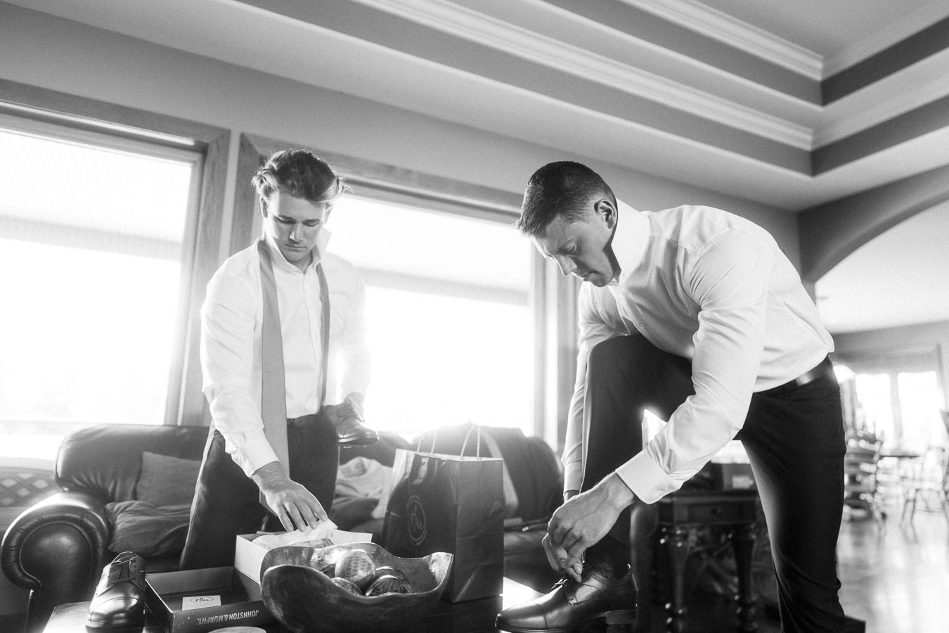 Two men in dress shirts preparing shoes beside a sofa during groom preparations at home