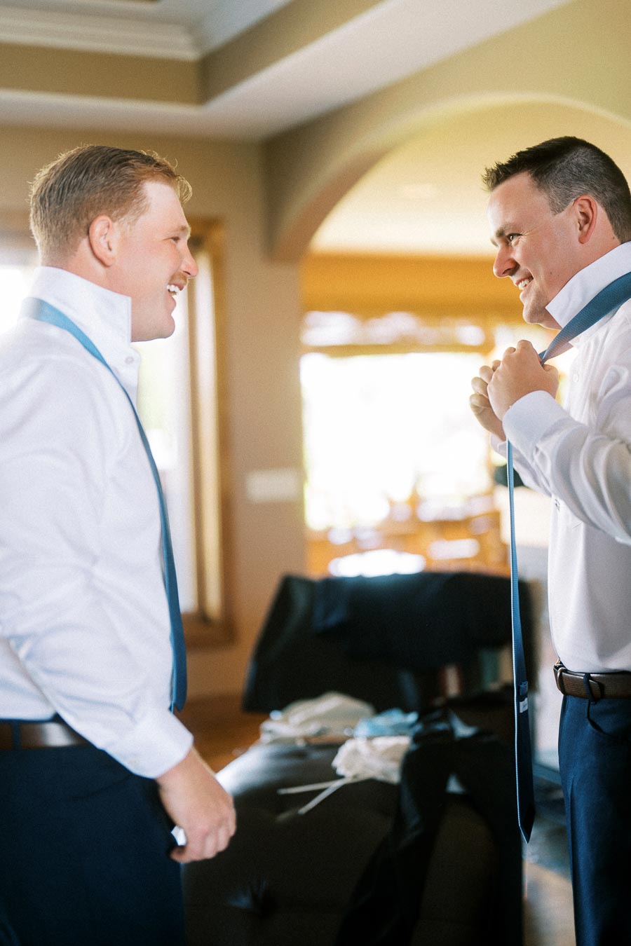 Two men in white shirts adjusting blue ties during an indoor groom preparation moment.
