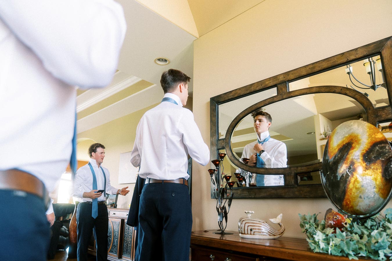 Groom adjusting blue tie in a mirror while friends get ready indoors.