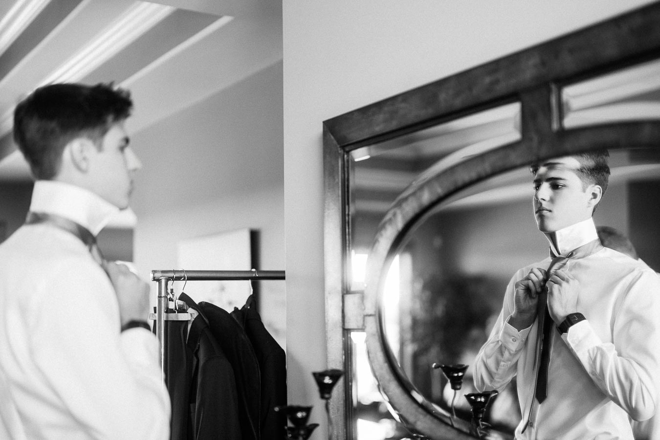 Groom adjusting his tie in a mirror during indoor wedding preparations.