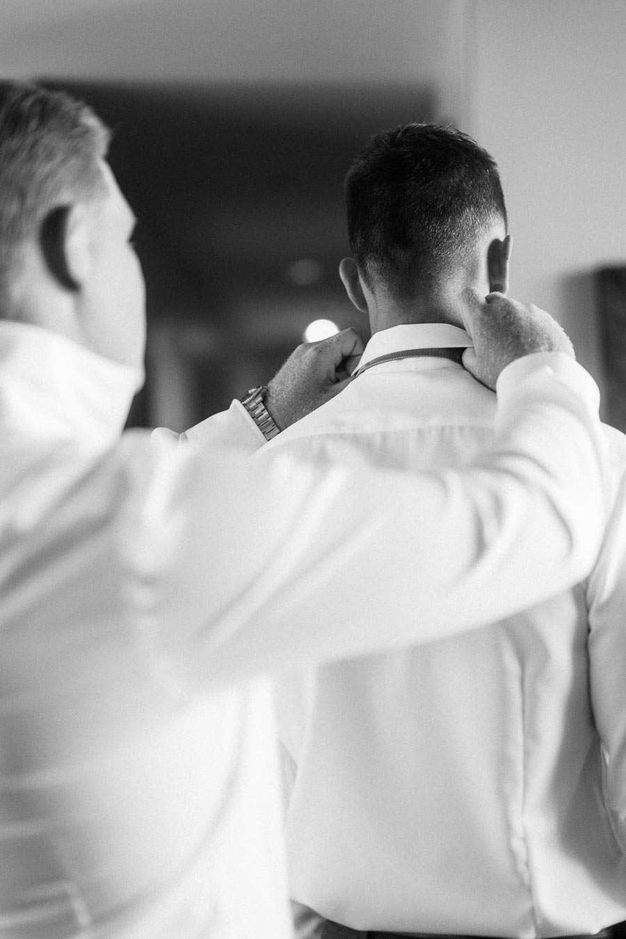 Man adjusts another man's collar during indoor wedding preparations in black and white.