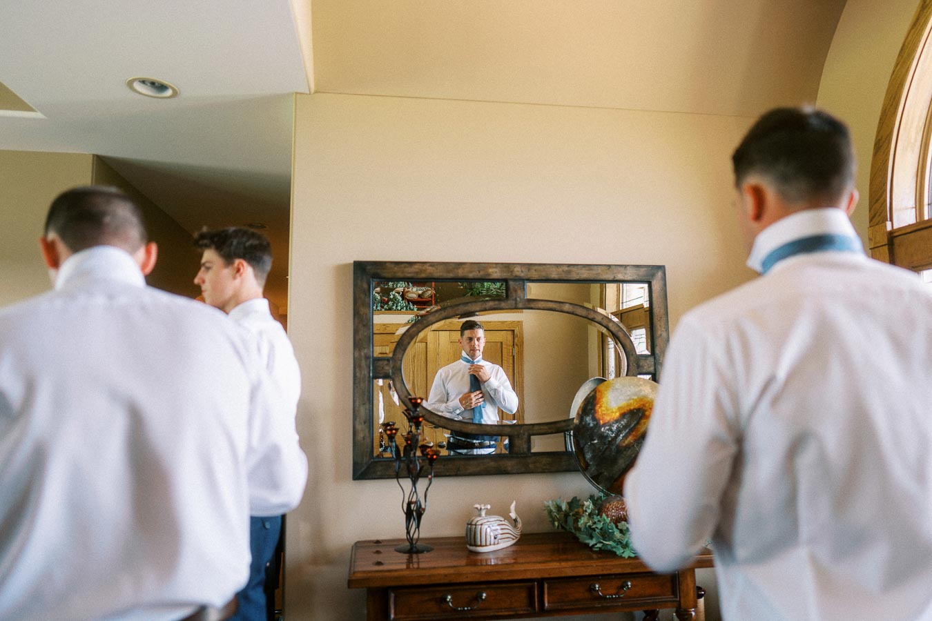 Groom and groomsmen in white shirts adjusting ties in a home mirror.