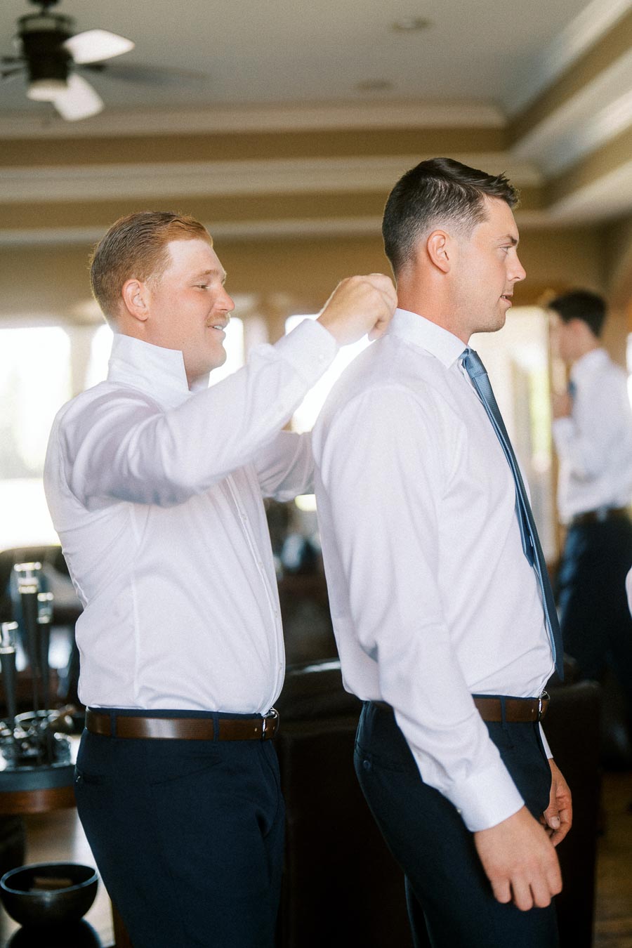 Groomsman adjusts a groom’s collar as they get ready indoors in white shirts and ties.