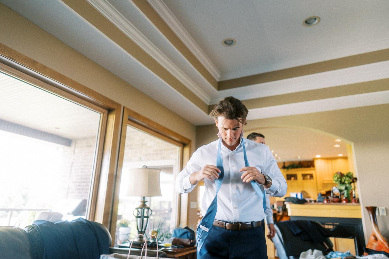 Groom adjusting a blue tie over a white shirt during wedding preparations at home.