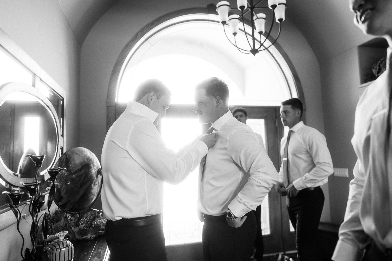 Groom and groomsmen adjusting ties in white shirts during indoor wedding preparations.