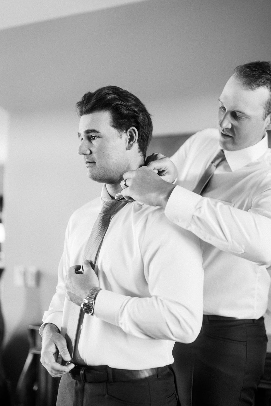 Man in a white shirt adjusts his tie while another man helps with the collar indoors.