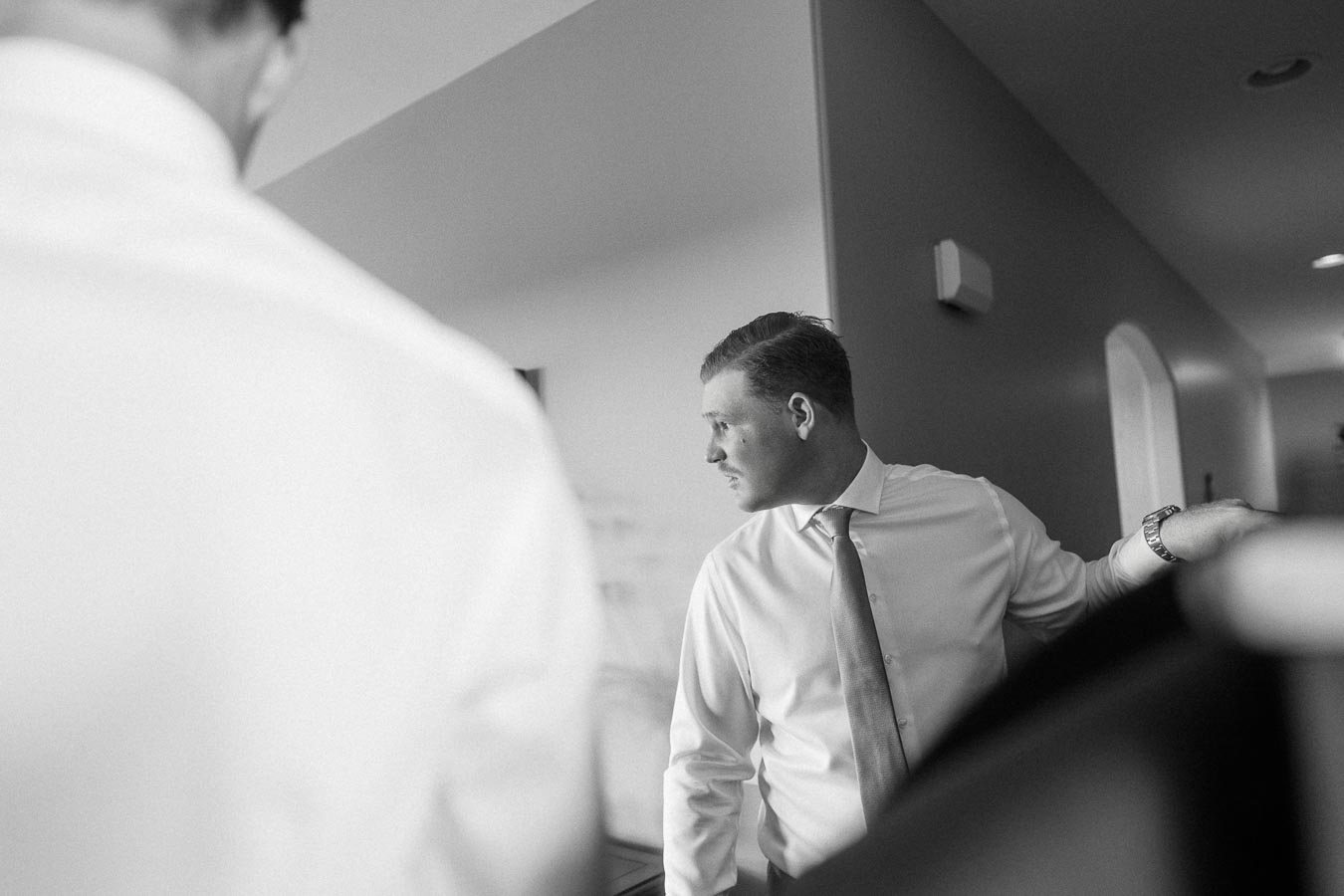 Groom in a dress shirt and tie stands indoors during wedding preparations.