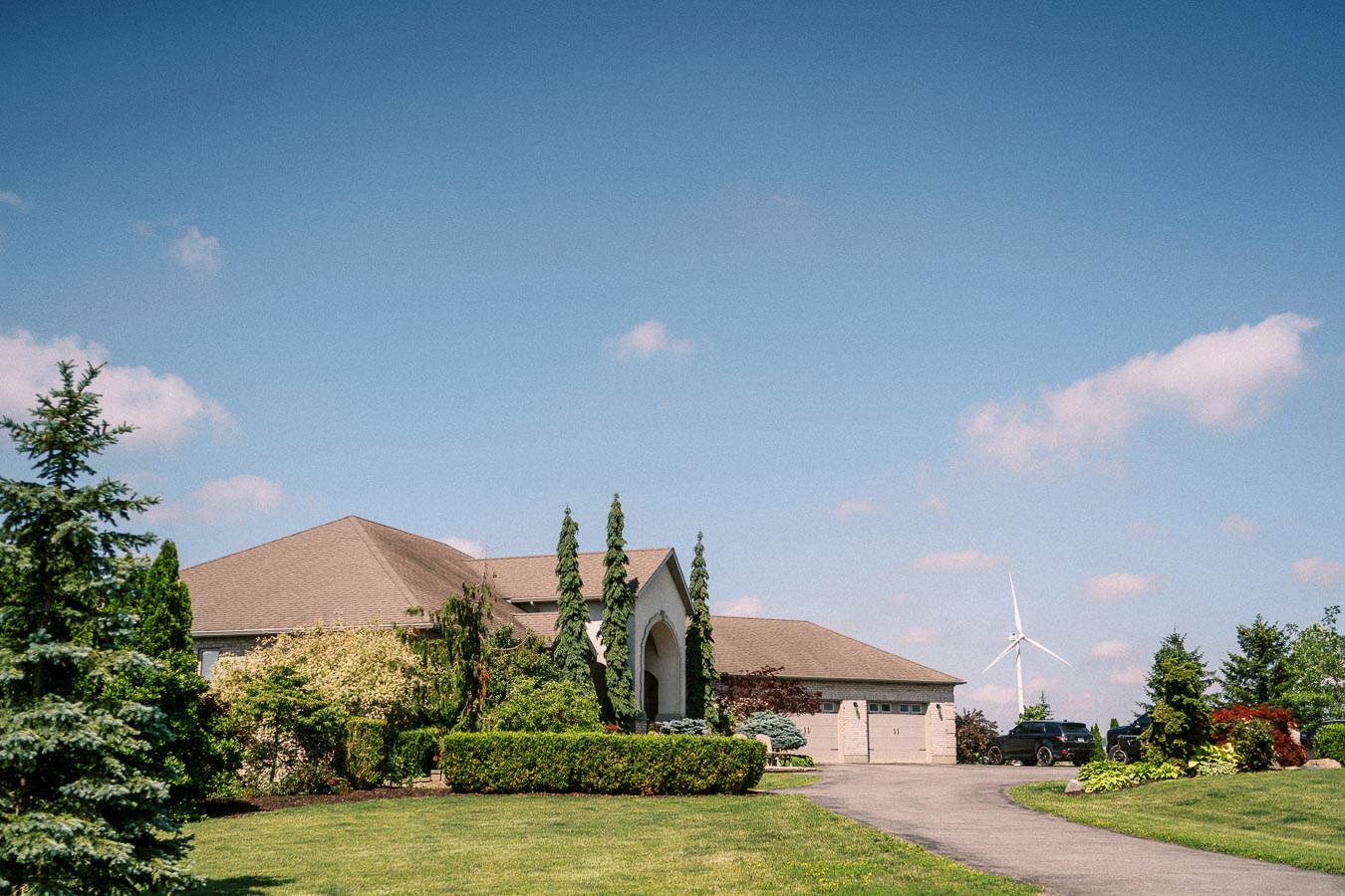 Large suburban home with manicured lawn, trees, driveway, and wind turbine under blue sky.