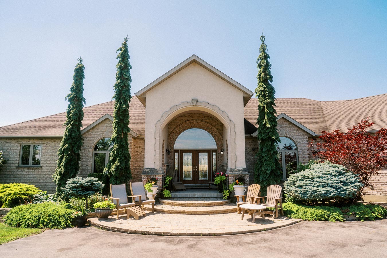 Large brick house entrance with arched doorway, patio chairs, shrubs, and tall evergreens.