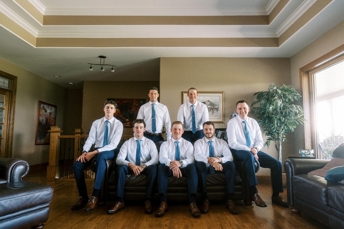 Seven men in white shirts and blue ties posing in a living room before a wedding.