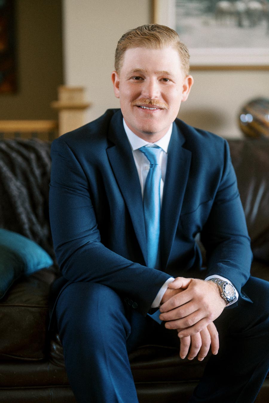 Groom in a navy suit and blue tie sitting on a leather sofa indoors before a wedding in Chatham-Kent, Ontario, Canada.