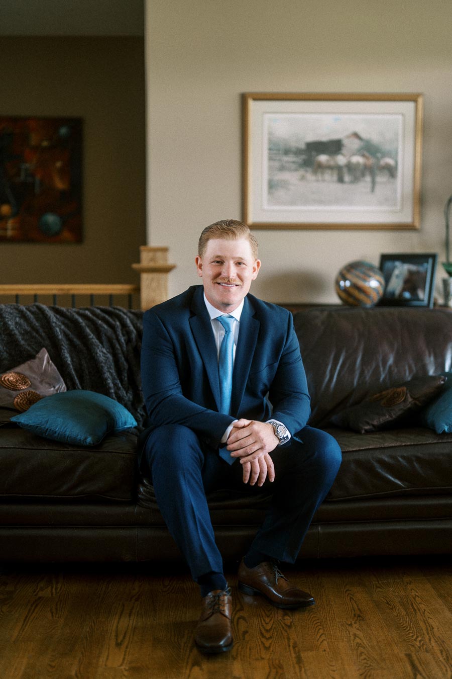 Smiling groom in a navy suit sits on a dark leather sofa inside a family home living room before a wedding.