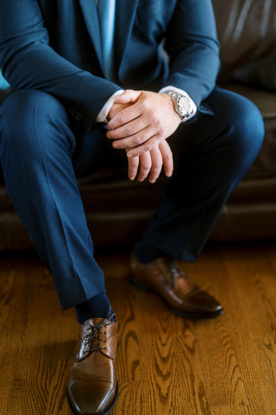 Seated groom in a navy suit with clasped hands, wristwatch, brown dress shoes, leather sofa, and wooden floor.