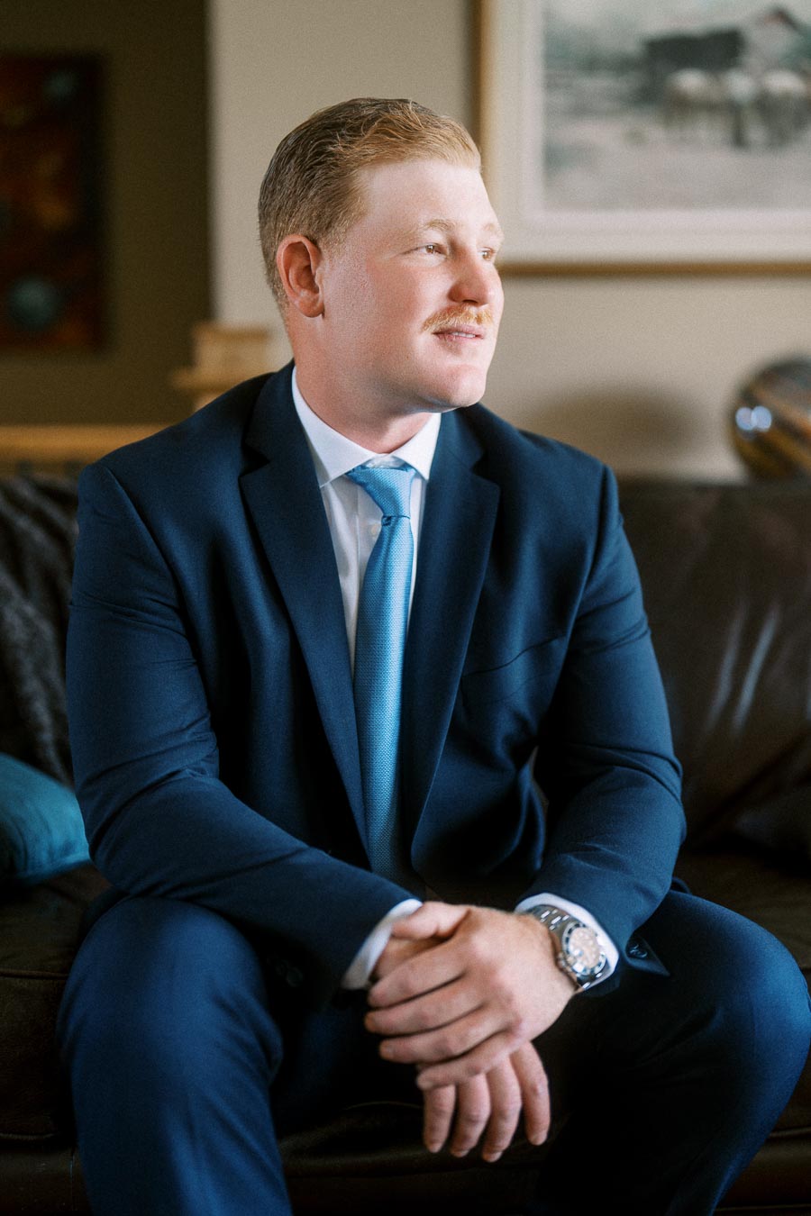 Groom in a navy suit with a light blue tie sitting on a leather sofa indoors before a wedding near Chatham-Kent, Ontario.