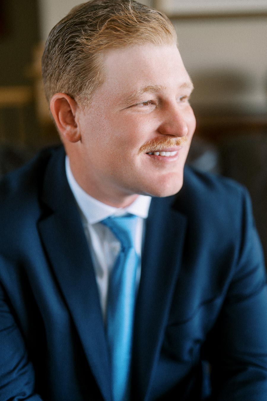 Smiling groom in a navy suit and blue tie looking toward window light during wedding preparations indoors.