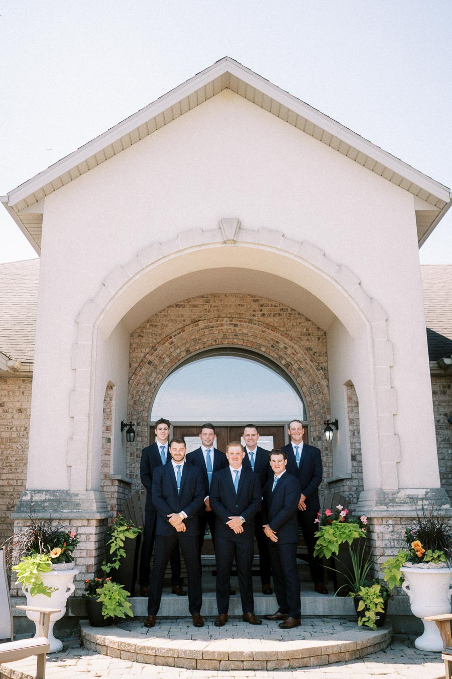 Seven men in dark suits pose on the steps of a brick house entrance with a large arched doorway and potted flowers.